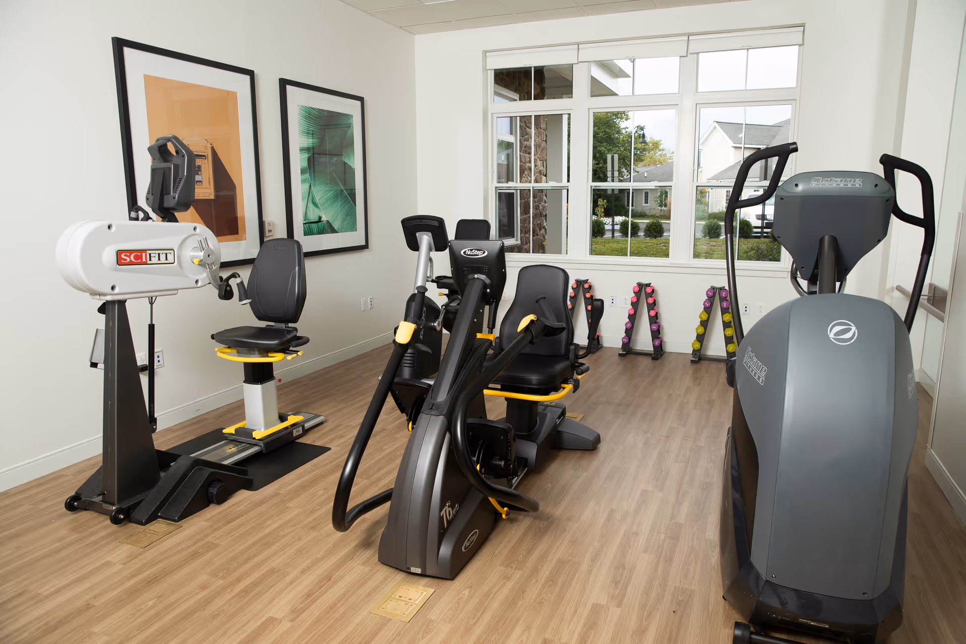 A small fitness room with exercise equipment including a SciFit recumbent bike, a NuStep machine, and a large elliptical trainer. There are racks of colorful dumbbells against the wall under a large window showing a view of the outside. The room has light wood flooring, white walls, and two framed pictures hanging on the left wall.