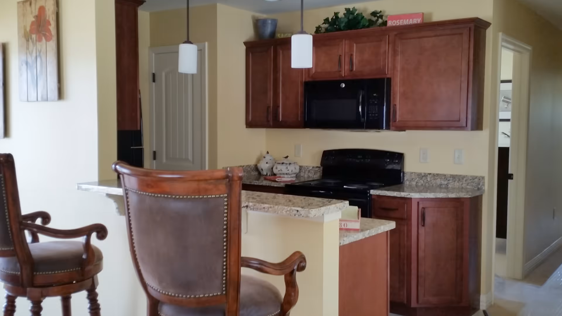Interior view of a kitchen area with wooden cabinets, a black microwave and stove, granite countertops, and two wooden bar stools with cushioned seats. There are two pendant lights hanging from the ceiling and a painting with red flowers on the wall.