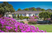 Single-story building framed by a manicured lawn and vibrant purple flowering shrubs with hills and a blue sky in the background.