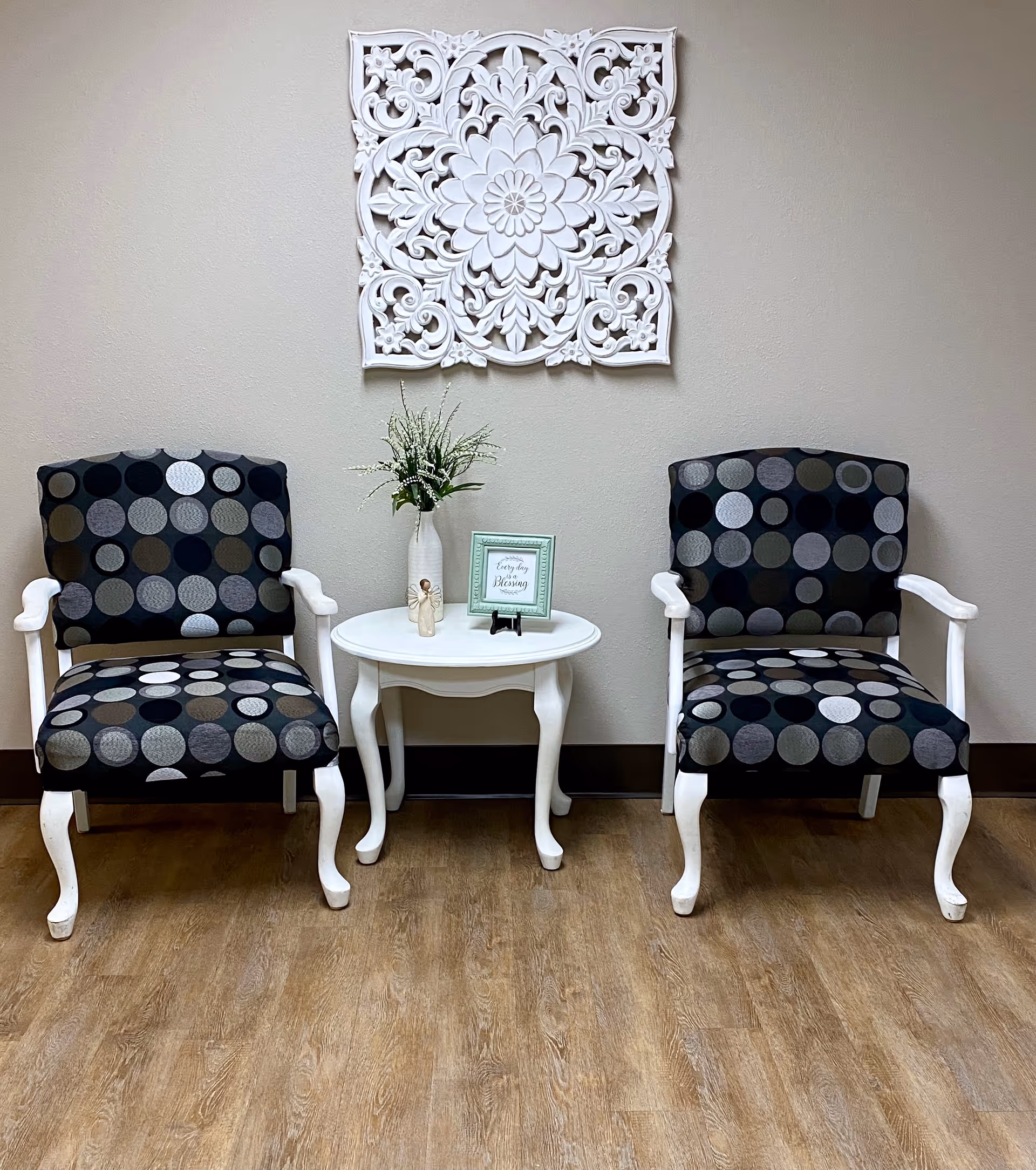 Two upholstered armchairs with a black and gray circular pattern flank a small white wooden table. On the table is a white vase with green foliage, a small angel figurine, and a framed sign. Above the table hangs an ornate white decorative wall piece. The setting is indoors with a wooden floor and beige walls.