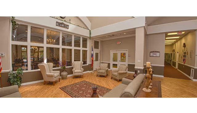 A comfortable senior living lounge with upholstered chairs and sofas arranged around a rug, large interior windows, and a hallway entrance.