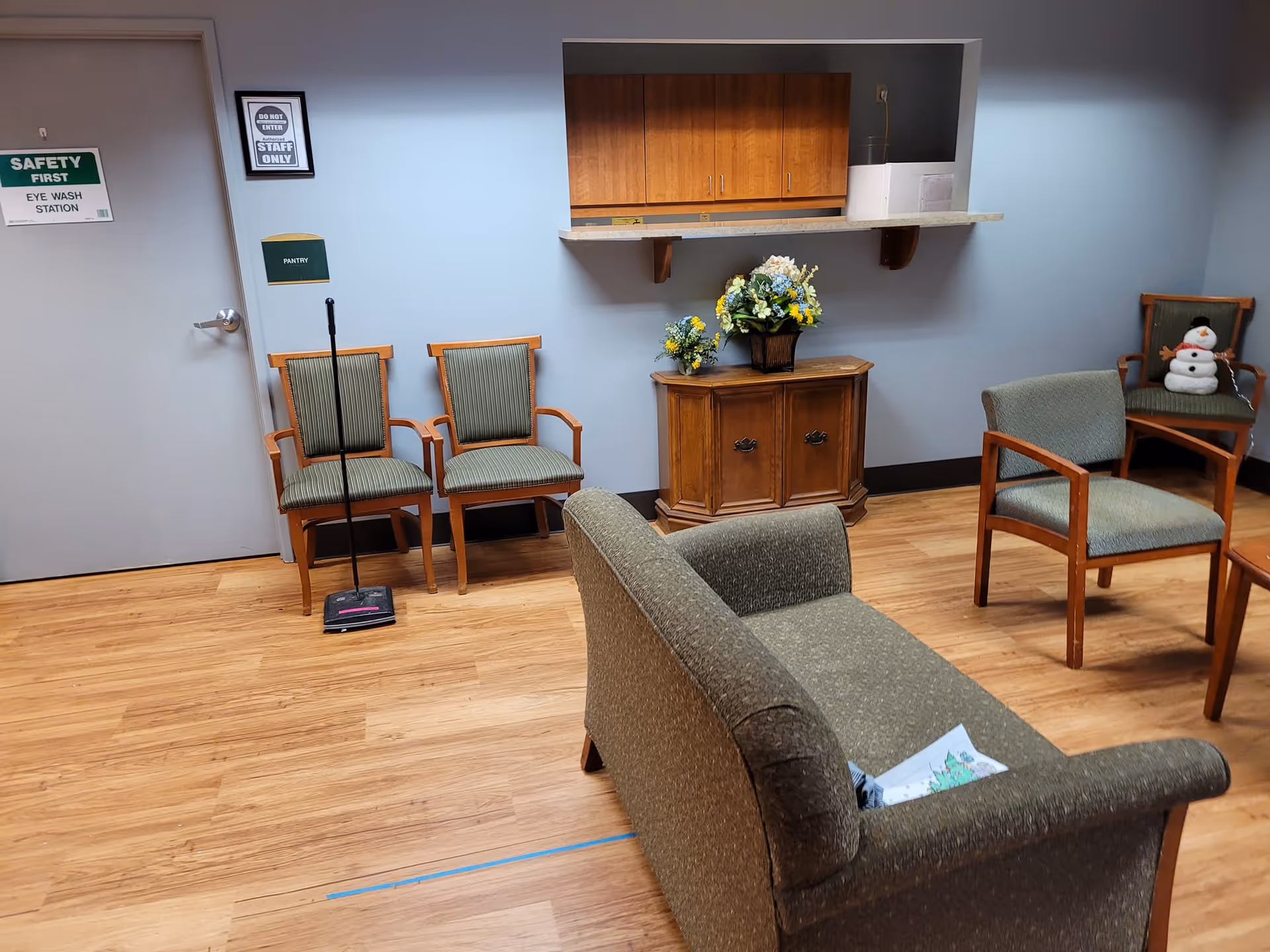 A small waiting or sitting area with wooden floor and light blue walls. There are several green cushioned chairs and a small wooden cabinet with flower arrangements on top. A door labeled 'Safety First Eye Wash Station' and a sign 'Do Not Enter Staff Only' are visible on the left side. A broom and dustpan lean against the wall near two chairs. A small snowman decoration sits on a chair in the back right corner.