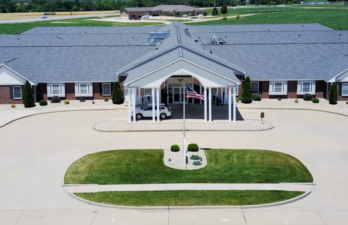 Front exterior view of Garden Place Waterloo facility showing a single-story building with a covered entrance, an American flag on a flagpole in front, a parked car under the entrance canopy, and landscaped greenery with bushes and grass.