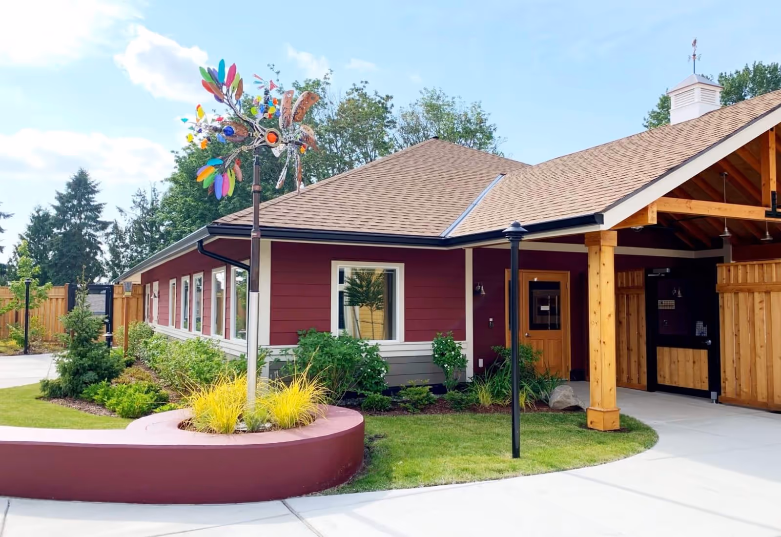 Exterior view of a single-story building with red siding and a brown shingled roof, surrounded by landscaped greenery and a curved planter with yellow ornamental grass. A colorful metal wind sculpture is mounted on a pole near the building. The entrance features wooden posts and a covered porch area.