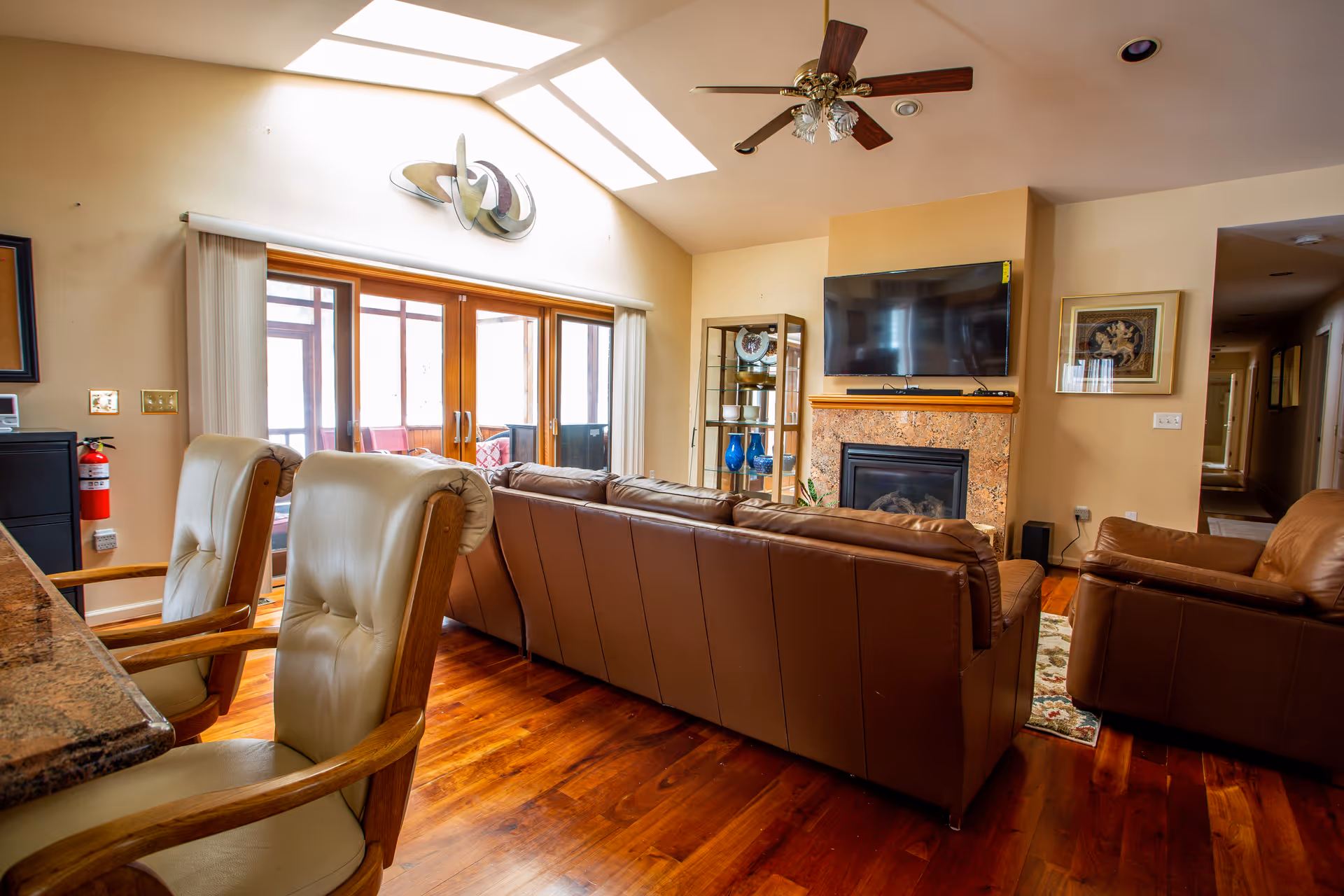 A cozy living room with a brown leather sofa and armchair facing a wall-mounted flat-screen TV above a fireplace. The room has wooden floors, a ceiling fan with lights, and large sliding glass doors with vertical blinds leading to a sunroom. There are two cushioned chairs with wooden arms near a granite countertop, and decorative items on a glass shelf next to the fireplace.