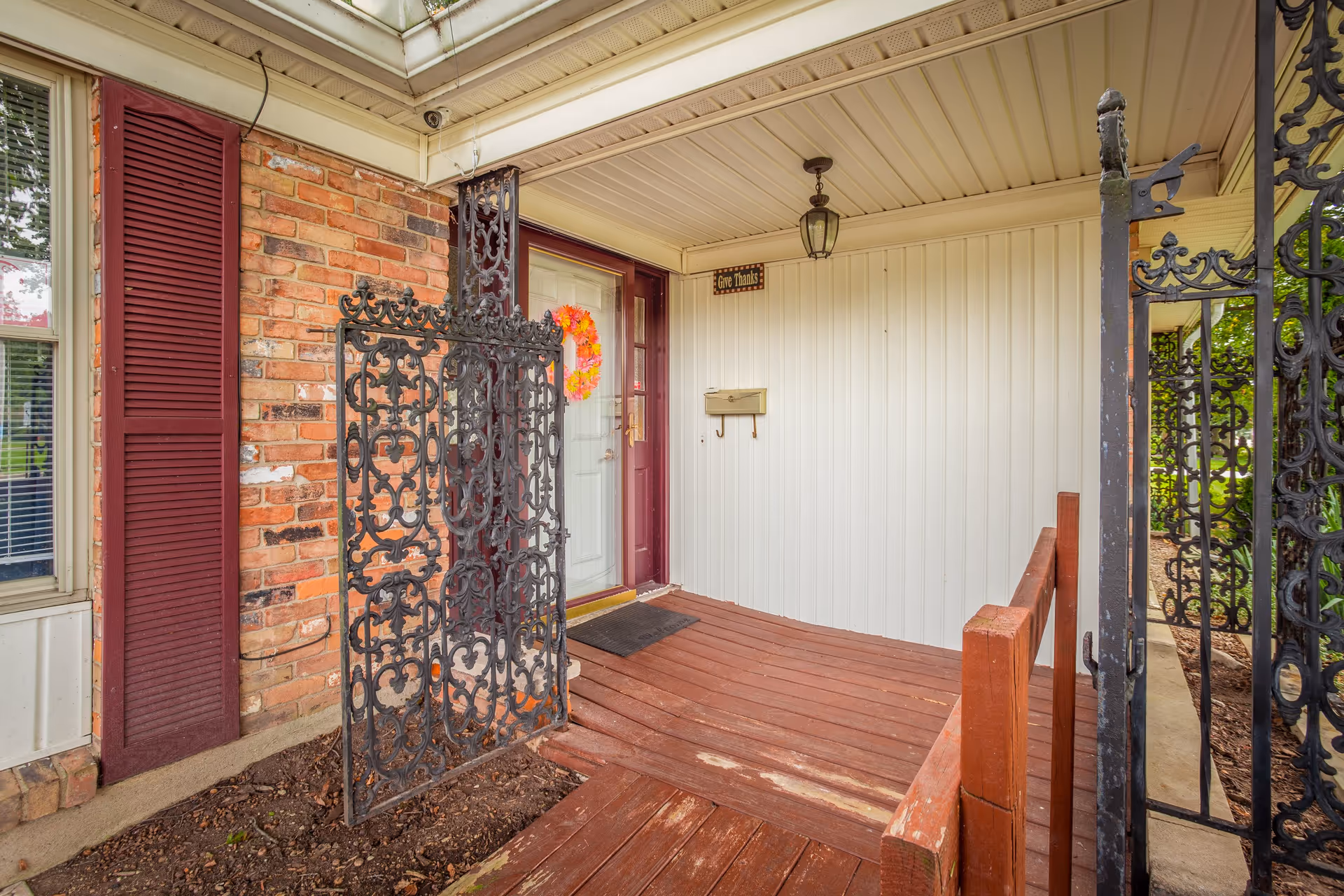 Front porch of a house with a wooden deck, decorative black wrought iron gate, brick wall with a maroon shutter, white door with a glass panel and a colorful wreath, a mailbox on the white siding wall, and a hanging lantern light. A small sign above the mailbox reads 'Give Thanks'.