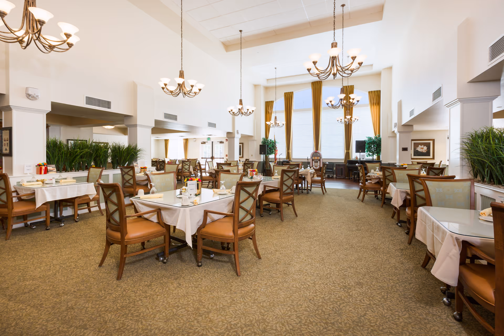 A spacious dining room in a senior living facility with multiple tables covered in white tablecloths, each set with plates, cups, and napkins. The room features high ceilings with several chandeliers, large windows with gold curtains, and green plants placed along the walls. The chairs have wooden frames with cushioned seats and backs.