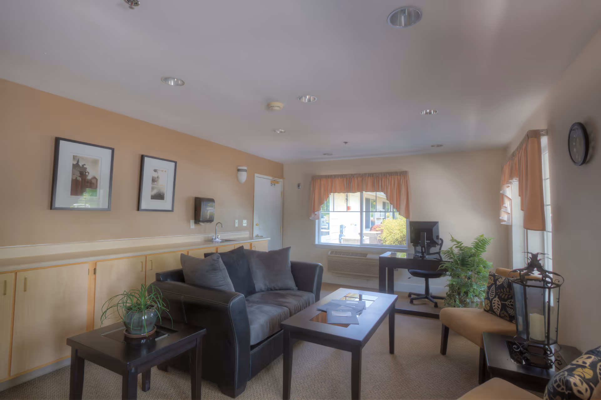 A cozy living room area with a dark gray sofa, two beige chairs with patterned cushions, a coffee table, and side tables. There is a small plant on one side table and a decorative lantern on the other. The room has beige walls with two framed pictures, a window with peach-colored curtains, a wall clock, and a desk with a computer near the window. Light fixtures are installed in the ceiling.