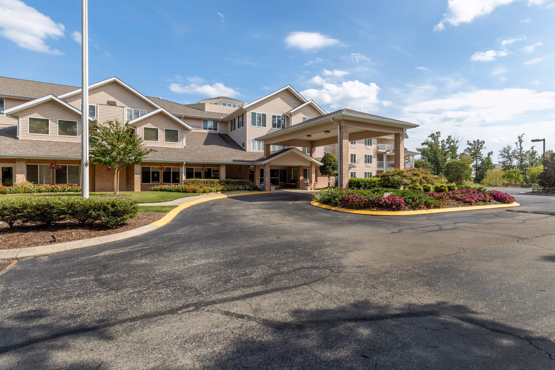 Exterior view of Creekside at Shallowford, a multi-story senior living community building with beige siding and brick accents. The entrance features a covered drop-off area with columns, surrounded by landscaped flower beds and shrubs. The sky is partly cloudy with blue patches.
