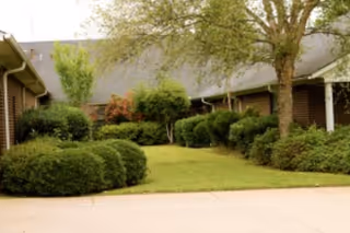 View of a landscaped outdoor area with trimmed bushes, trees, and grass in front of a building with a sloped roof and brick walls.