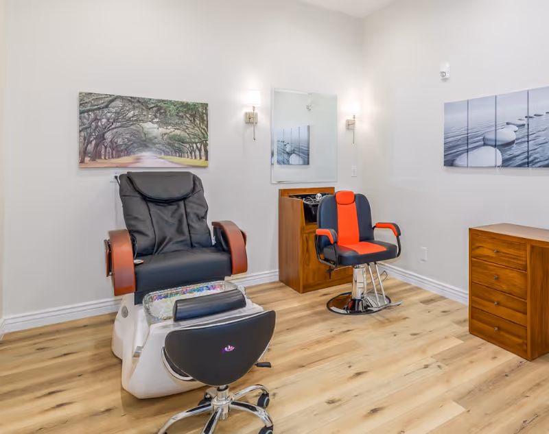 A clean and modern salon room with a black and brown pedicure chair, a black and red styling chair, a small wooden cabinet, a mirror on the wall, and two framed pictures of nature scenes. The floor is light wood, and the walls are white with two wall-mounted lights.