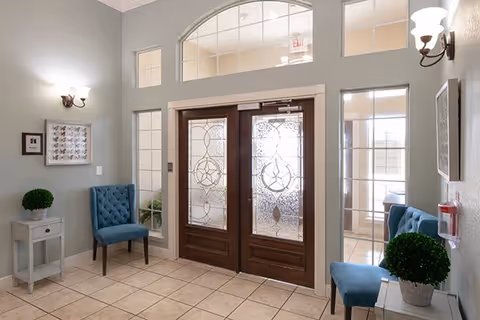 Entrance area with double wooden doors featuring decorative frosted glass panels. On each side of the doors are tall windows with multiple panes. The room has beige tiled flooring, light gray walls, and two blue upholstered chairs placed against the walls. Small white side tables with green potted plants are next to each chair. Wall sconces provide lighting, and there is a framed picture on the left wall.