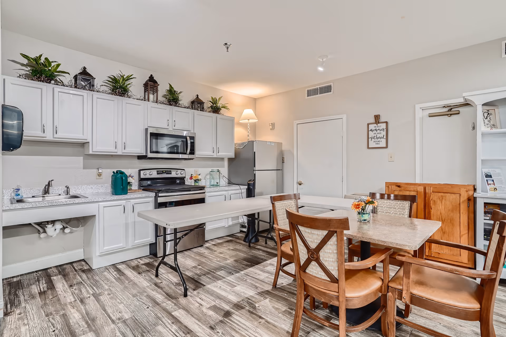 A kitchen and dining area with white cabinets, a stainless steel stove and microwave, a refrigerator, and a sink. There is a long folding table and a dining table with four wooden chairs. The floor has a wood-like pattern, and there are decorative plants and lanterns on top of the cabinets. A small flower arrangement is on the dining table.