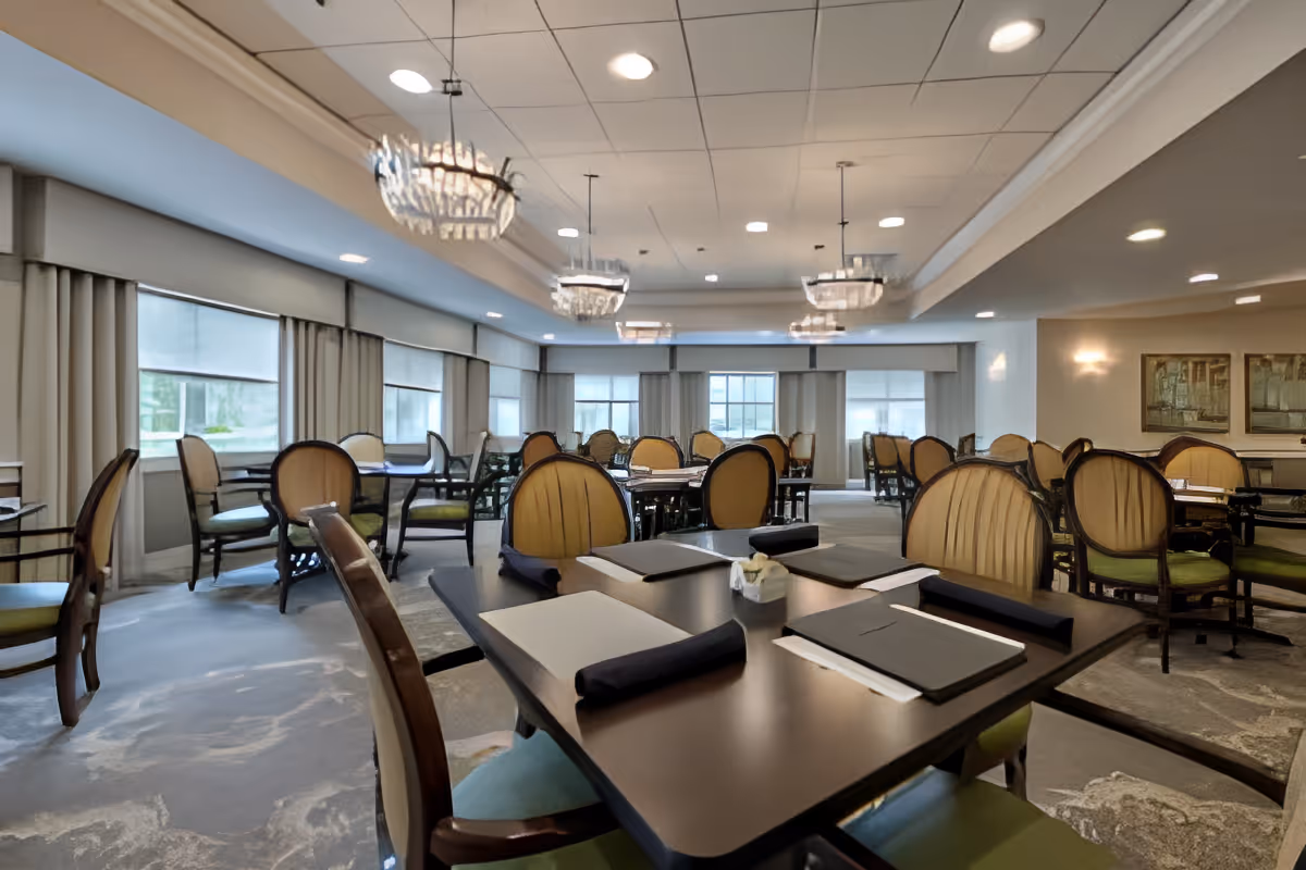 Spacious dining room with multiple set tables and upholstered chairs under chandeliers and large windows.