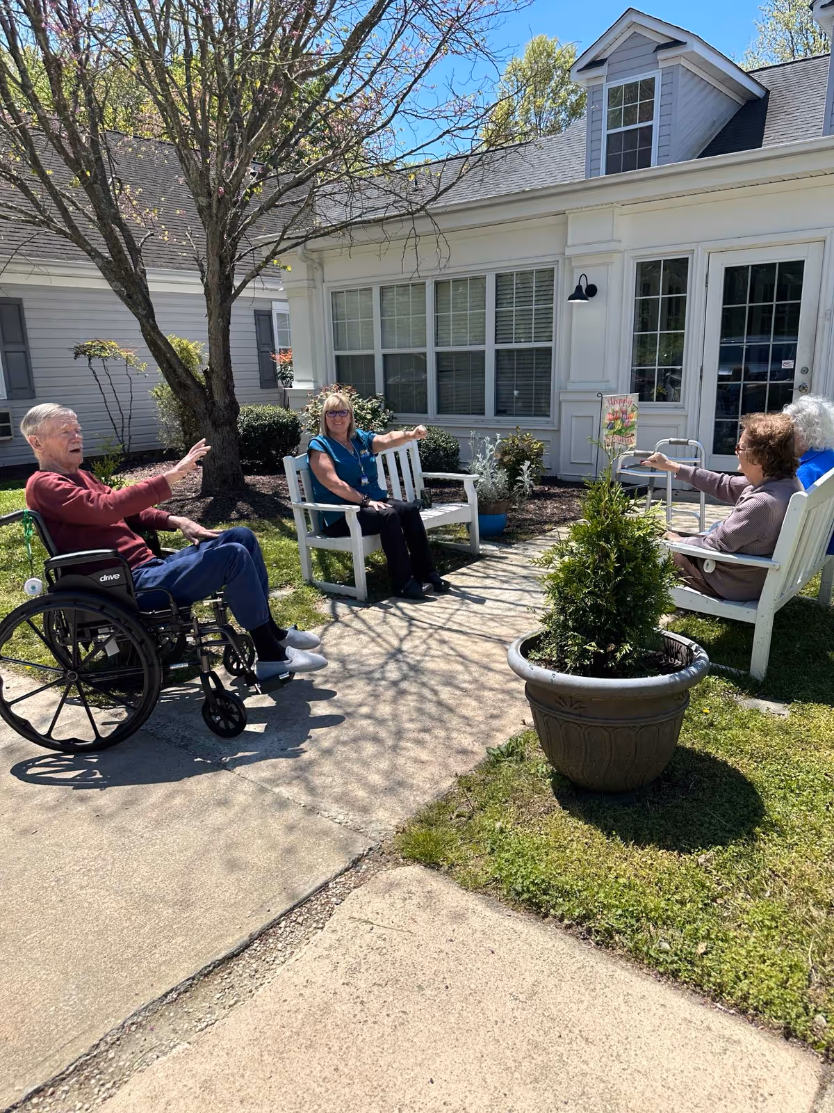 Four elderly people sitting outside in a garden area near a white building. One man in a wheelchair is on the left, two women are sitting on white benches, and another woman is seated on a white chair. They appear to be engaged in conversation on a sunny day with trees and plants around.