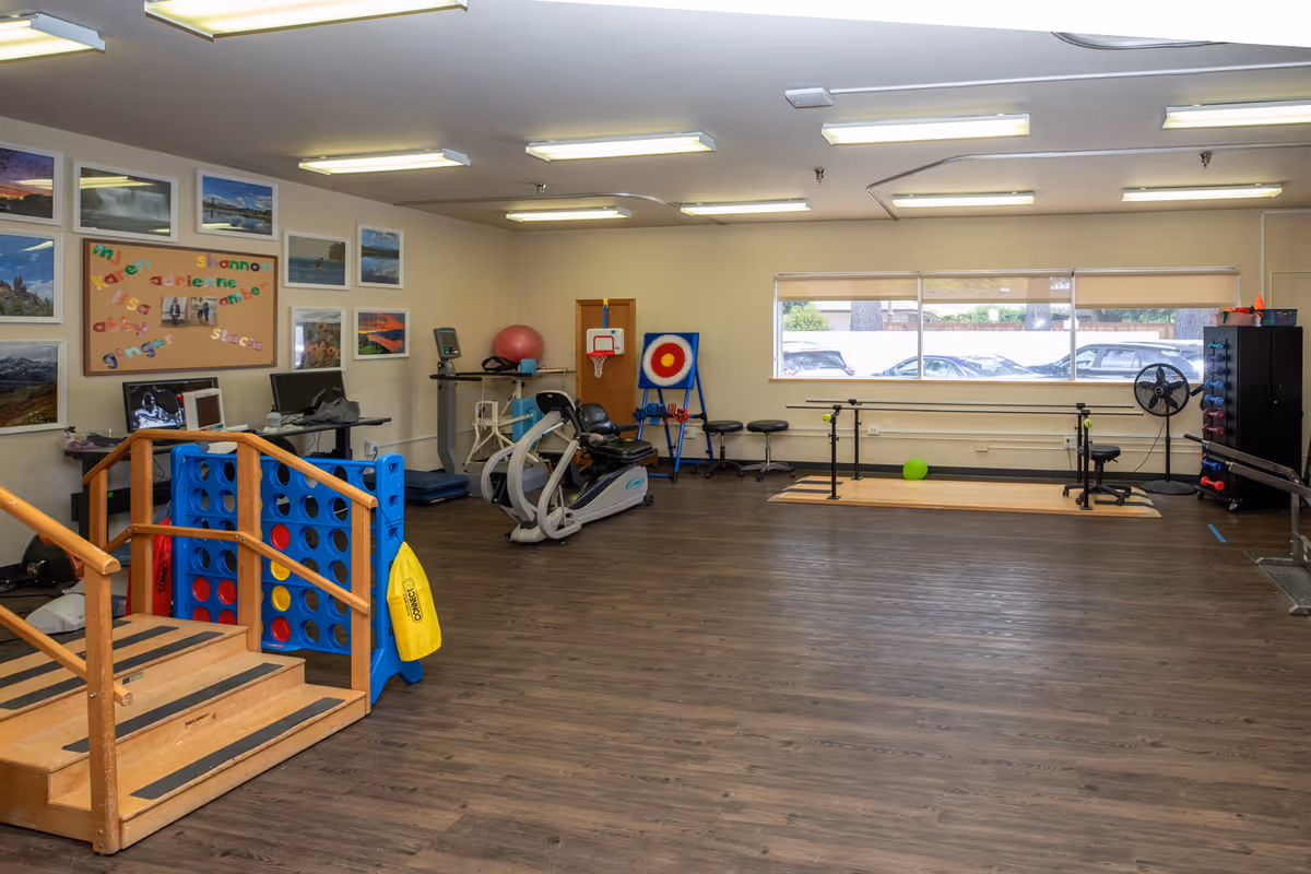 A spacious therapy or exercise room with wooden flooring, equipped with parallel bars for walking exercises, a recumbent exercise bike, a large Connect Four game, a small basketball hoop, and various therapy and exercise equipment. The room has a large window letting in natural light and several framed pictures on the walls.