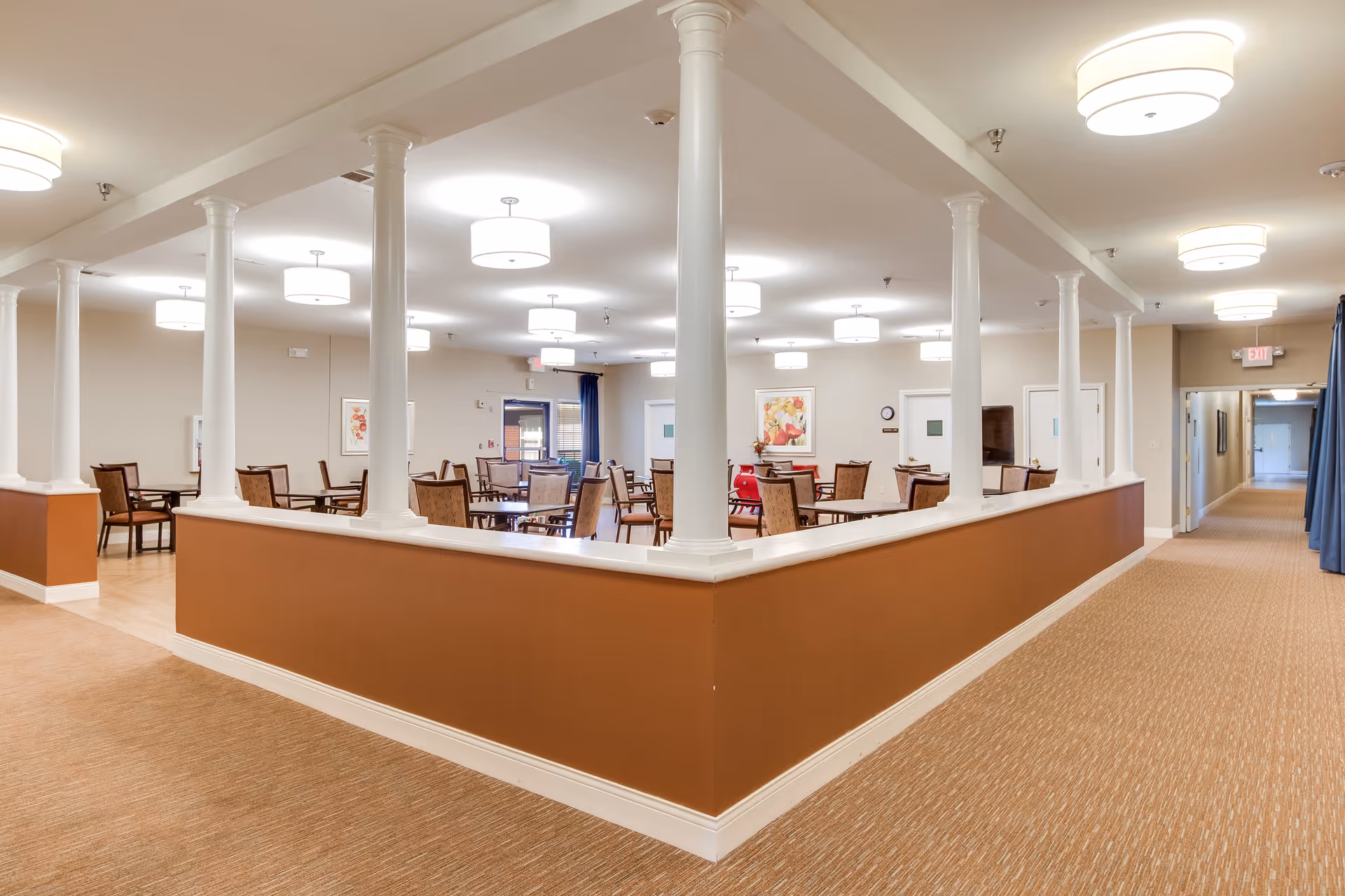 Open communal dining area with white columns, round tables and chairs under ceiling lights next to a long hallway.