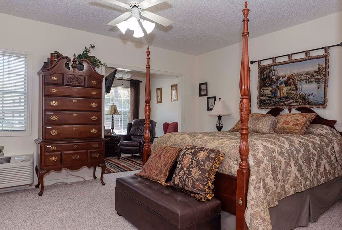 A traditional bedroom featuring a large wooden four-poster bed with patterned bedding and decorative pillows. At the foot of the bed is a dark brown leather bench. To the left, there is a tall wooden dresser with multiple drawers and decorative brass handles. The room has beige walls, carpeted floor, and a ceiling fan with lights. In the background, an adjoining sitting area is visible with a leather armchair, a lamp, and a window with curtains.