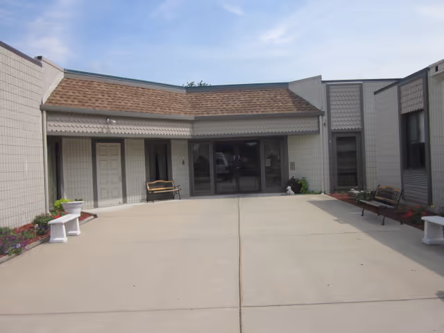 Exterior view of a single-story building with a concrete courtyard in front. The building has light-colored walls, a brown shingled roof, and multiple windows. There are benches and flower beds along the sides of the courtyard.