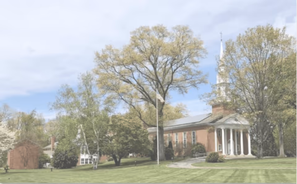 A large brick building with white columns and a tall steeple, surrounded by green trees and a well-maintained lawn under a partly cloudy sky.