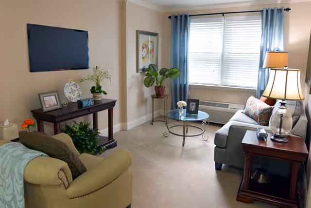 Bright living room with a sofa, armchair, glass coffee table, side tables with lamps, wall-mounted TV, and a window with blue curtains.
