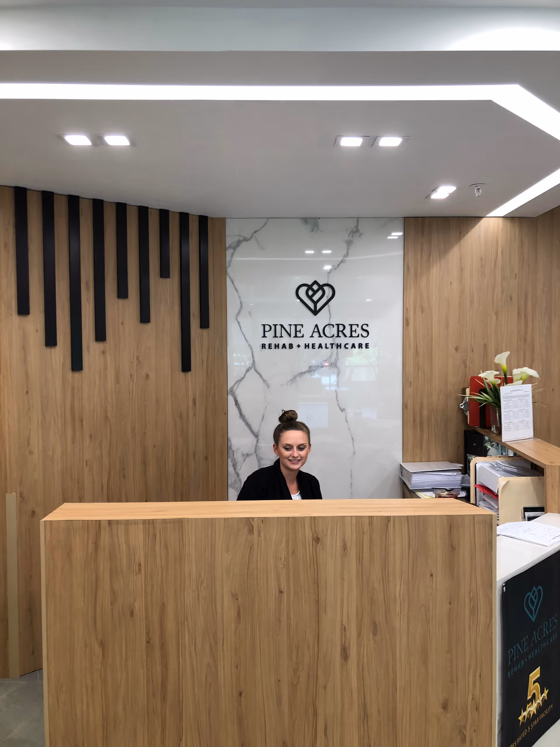 Reception desk at Pine Acres Rehab & Healthcare with a smiling receptionist in front of a wood-paneled wall and facility logo.