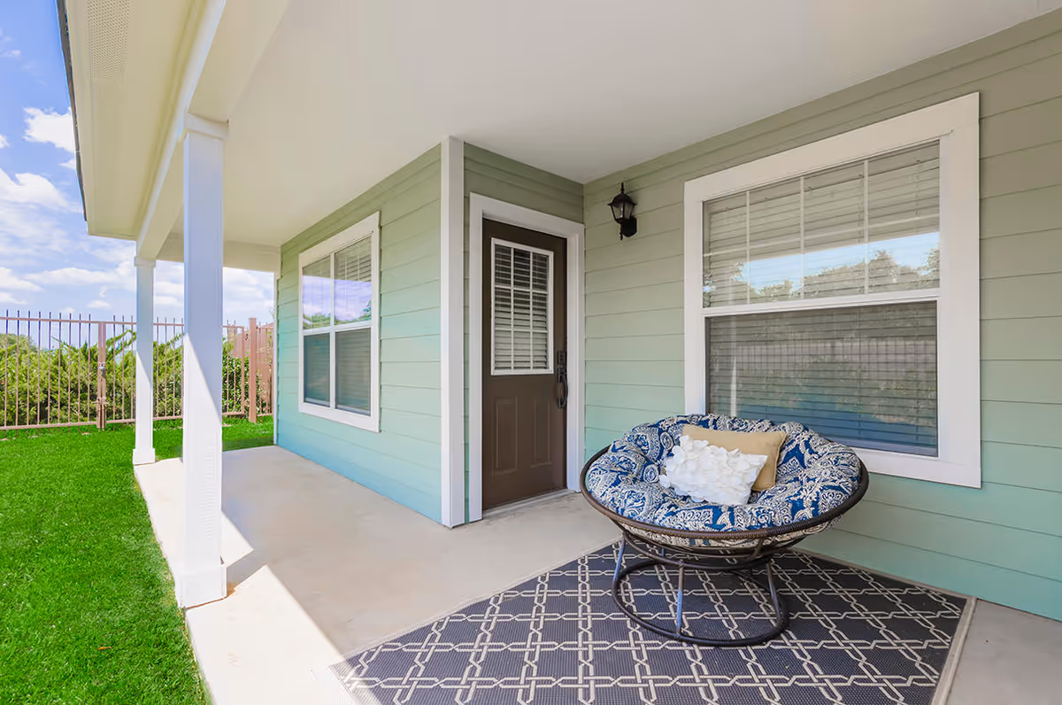 Covered outdoor patio area with a cushioned round chair on a patterned rug, a brown door with a window and white blinds, two windows with white frames and blinds, light green siding on the building, white pillars supporting the roof, and a green lawn with a metal fence in the background under a partly cloudy sky.