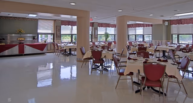 A spacious dining room with multiple tables covered in white tablecloths and set with plates, glasses, and napkins. The room has large windows with red valances allowing natural light to fill the space. There are red cushioned chairs around the tables and a buffet area with a red and white tablecloth along one wall.