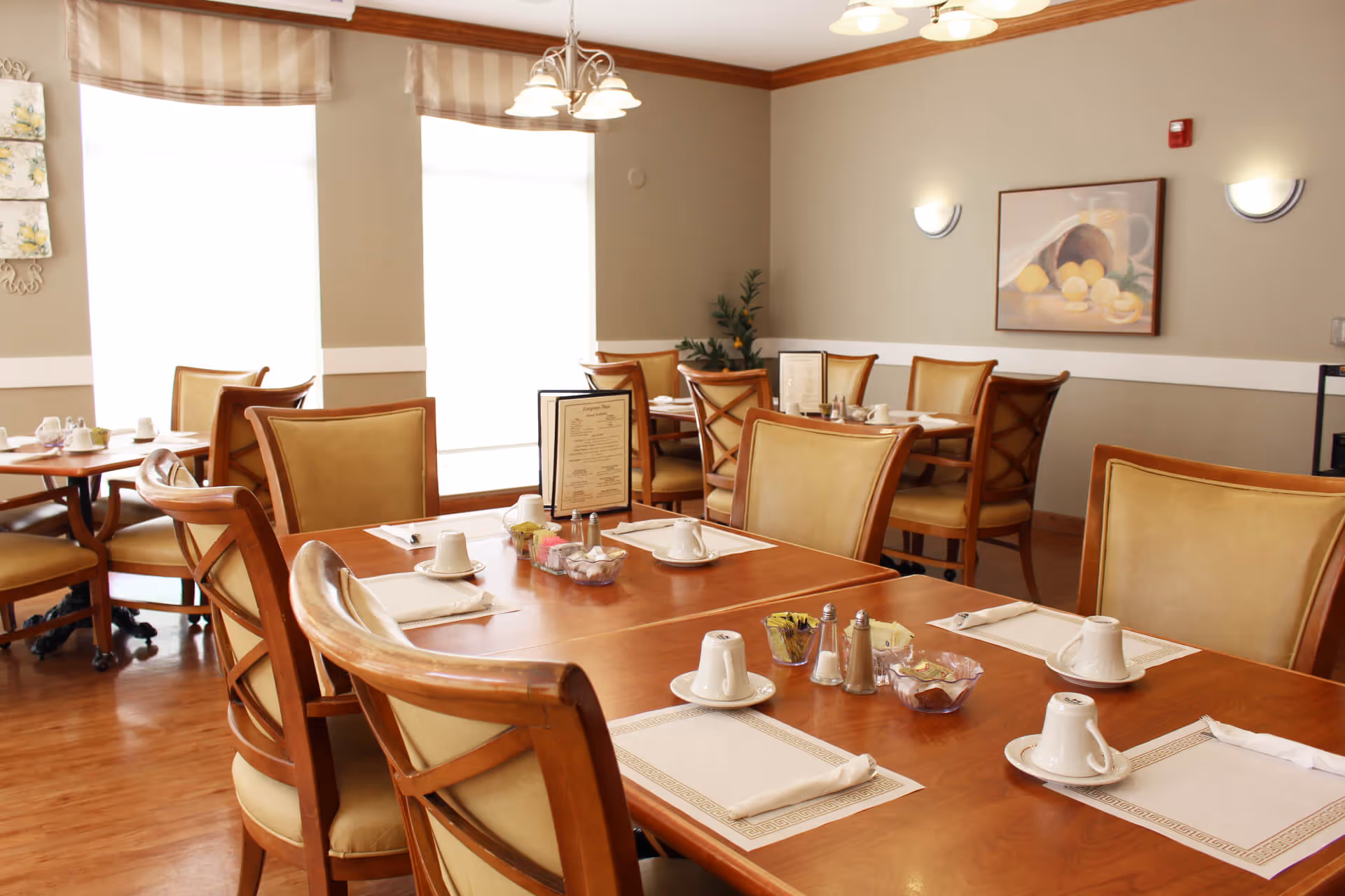 A well-lit dining room with wooden tables and cushioned chairs arranged neatly. Each table is set with white cups, saucers, napkins, salt and pepper shakers, and small bowls containing sugar packets. The room has large windows with striped valances, beige walls with white trim, wall-mounted lights, and a framed painting of fruit on the wall.