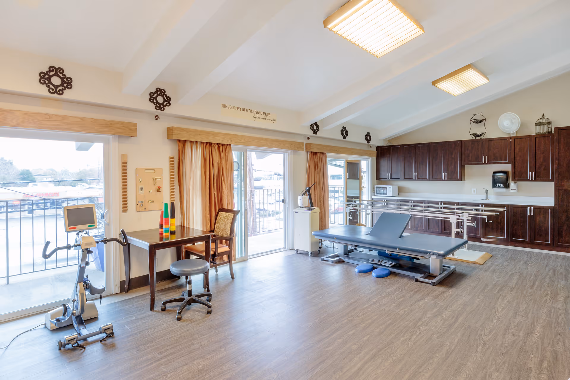 Bright rehabilitation/therapy room with an adjustable treatment table, parallel bars, exercise bike, table and chairs, and large sliding glass doors to a balcony.