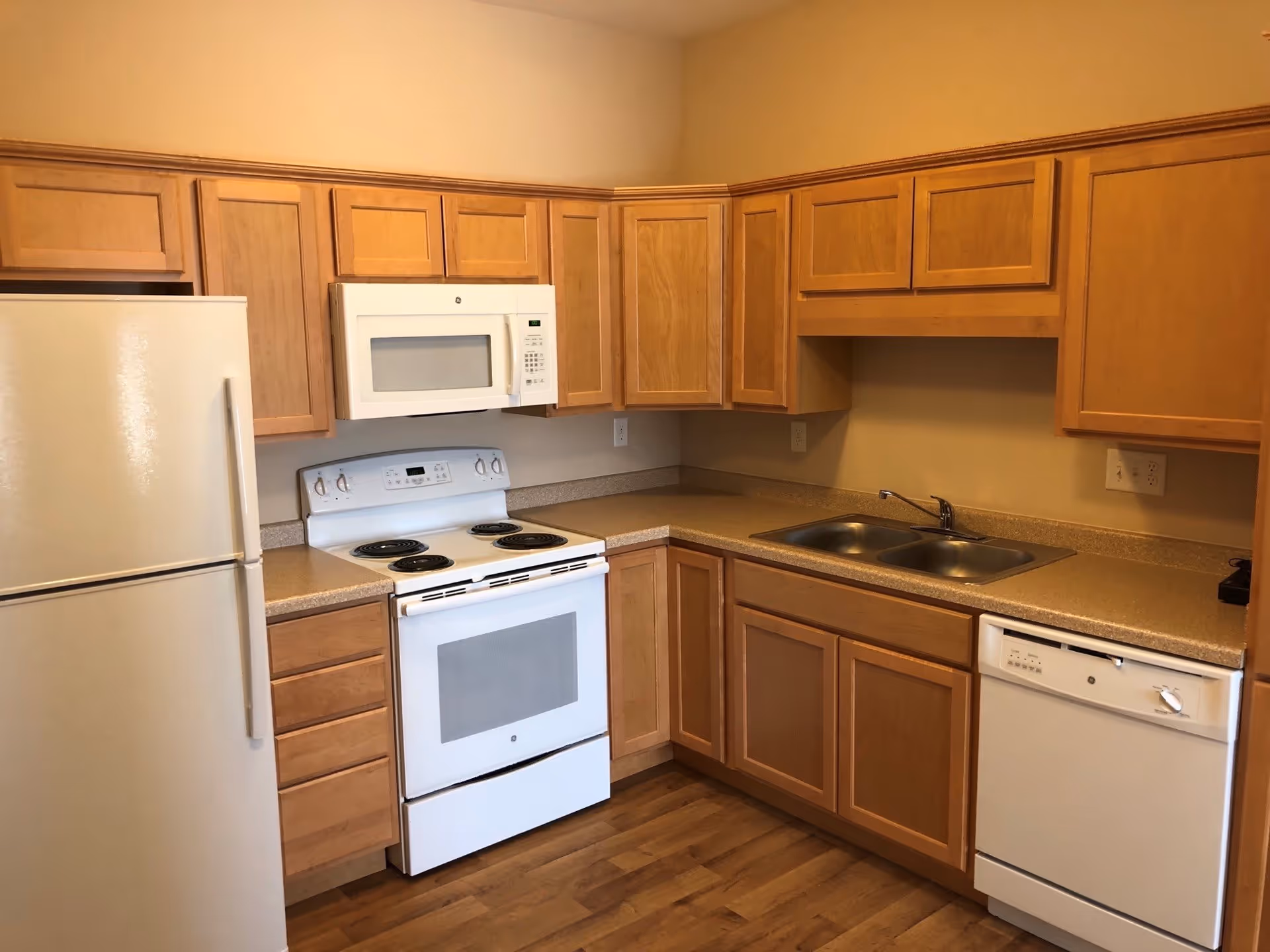 A kitchen with wooden cabinets, a white refrigerator, a white electric stove with an oven, a white microwave above the stove, a double stainless steel sink, and a white dishwasher. The countertops are beige, and the floor is wooden.