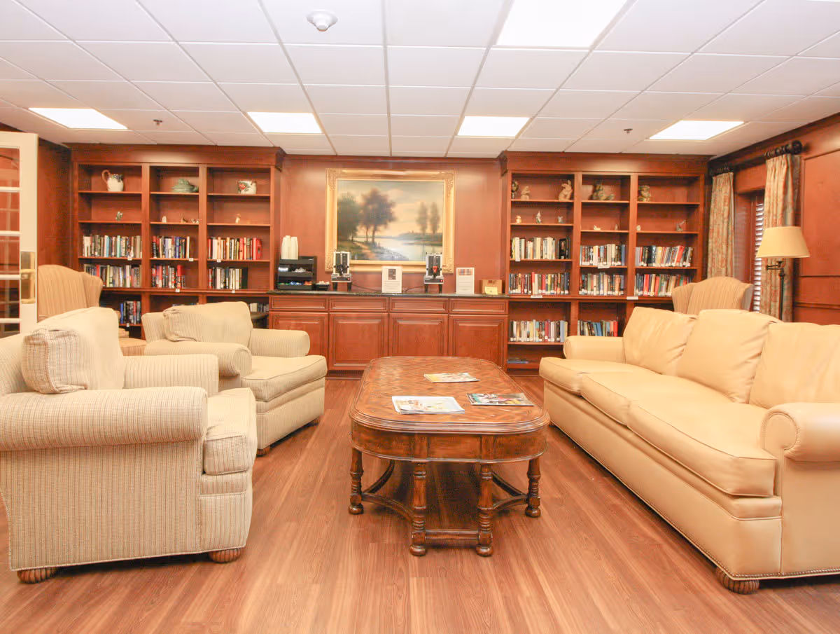A cozy living room area with a beige leather sofa, two beige armchairs, a wooden coffee table with magazines, and built-in wooden bookshelves filled with books and decorative items. A framed landscape painting hangs on the wood-paneled wall above a cabinet with coffee dispensers and cups.