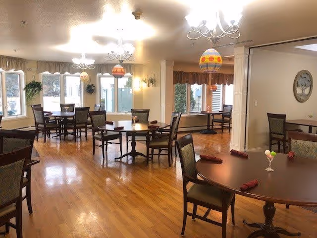A bright dining room with several round wooden tables and chairs arranged neatly. Each table has folded red napkins and a small centerpiece. Large windows allow natural light to fill the room, and decorative hanging lights with colorful patterns are suspended from the ceiling. The floor is polished wood, and the walls are painted a light color with some framed artwork and plants near the windows.