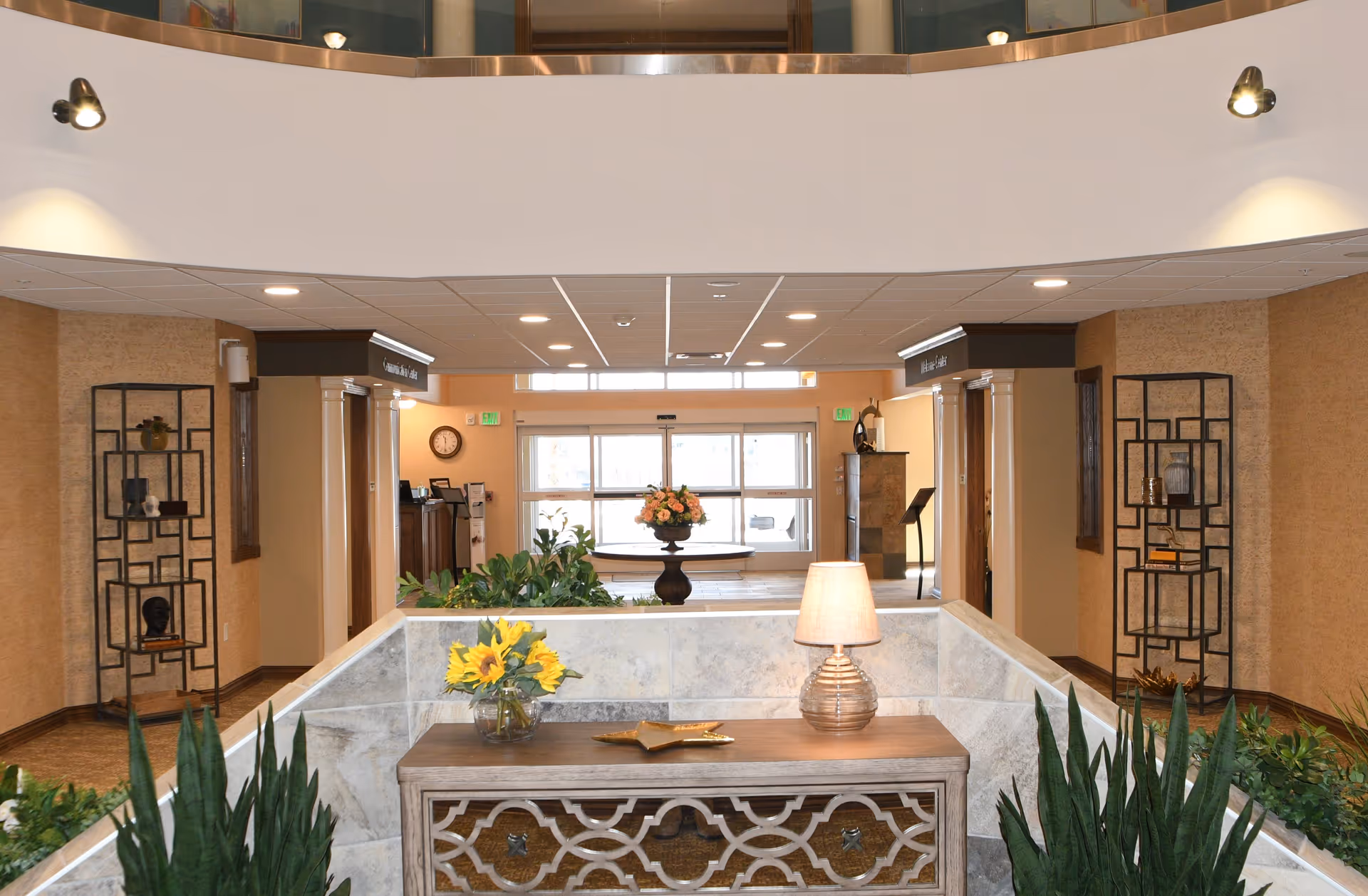 Interior view of a retirement community lobby with a decorative wooden console table holding a lamp and a vase of sunflowers. The space features plants, two black metal shelving units with decorative items, and a round table with a flower arrangement near the glass entrance doors. The ceiling has recessed lighting and the walls are painted in warm tones.