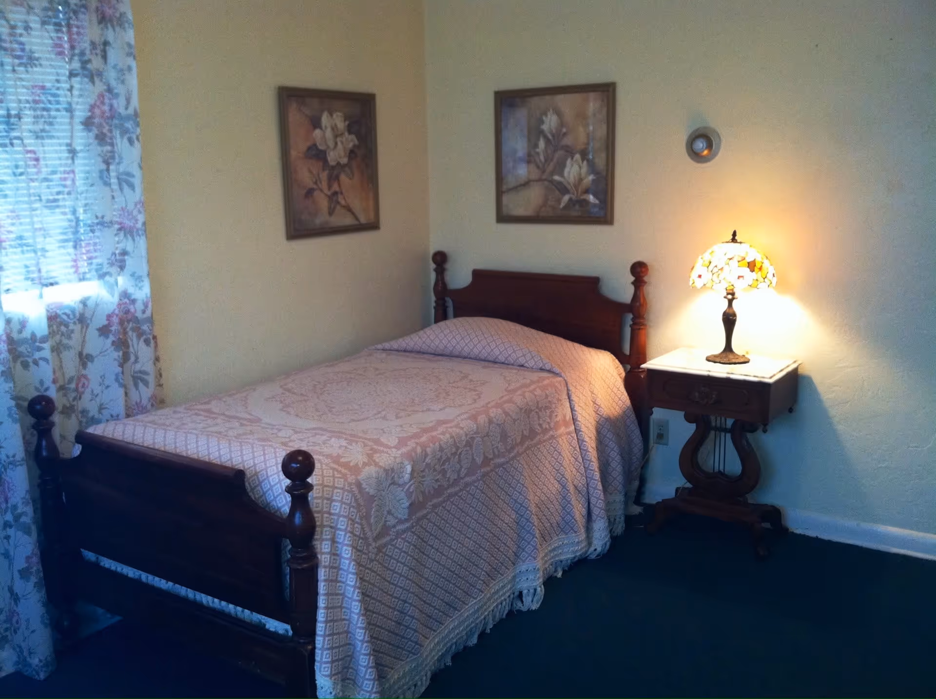 A small bedroom with a single wooden bed covered with a patterned bedspread. There is a wooden nightstand with a decorative lamp emitting warm light. Two framed floral paintings hang on the wall above the bed. A window with floral curtains is on the left side of the room.