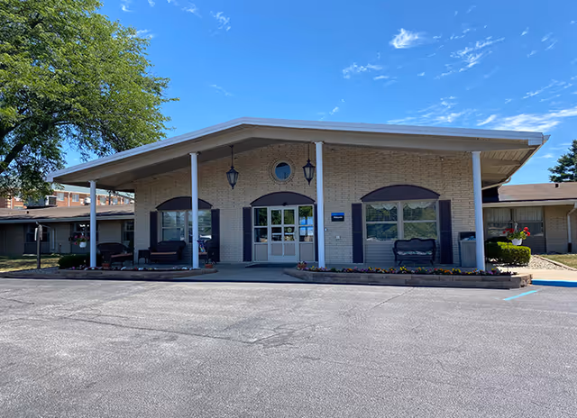 Front entrance of a low-rise brick care facility with a covered portico, benches, and a parking area.