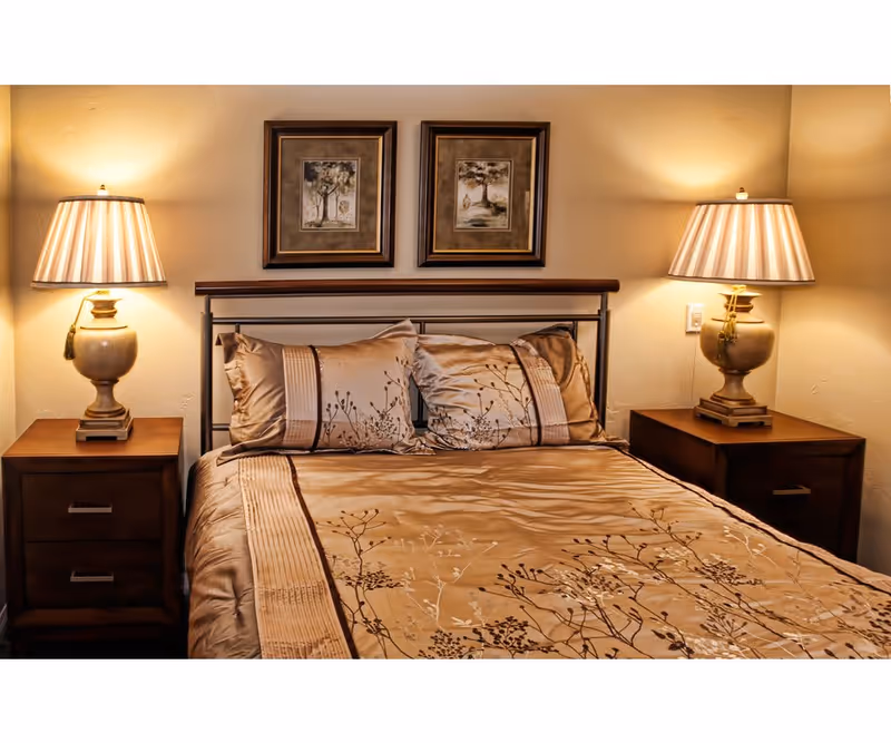 A neatly made bed with a beige and brown floral patterned comforter and matching pillows. The bed has a metal headboard and is flanked by two wooden nightstands, each with a lamp featuring a beige base and striped lampshade. Above the bed are two framed pictures with tree designs. The room has warm lighting and beige walls.