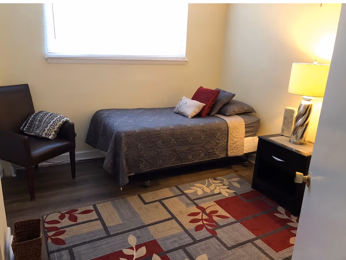 A small bedroom with a single bed covered in a gray patterned bedspread and several pillows. There is a dark wooden nightstand with a lamp on it next to the bed. A dark brown armchair with a folded blanket on it is placed near the window. The floor has a colorful area rug with red, beige, and gray geometric and leaf patterns. The room has light-colored walls and a window with blinds.