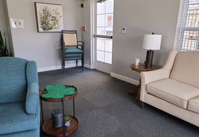 A small seating area with a blue armchair on the left and a beige sofa on the right. Between them is a round wooden side table with a lamp and a small decorative item. Another small round table with a green decorative piece and a small plant is in front of the blue armchair. In the background, there is a stack of chairs against a light gray wall with a framed floral artwork above them. A glass door and a window with blinds are visible on the right side.