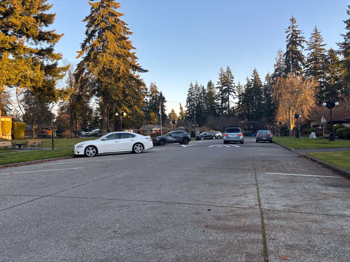 Parking lot with several cars, tall evergreen trees, grassy lawns and a clear sky at a senior living campus.