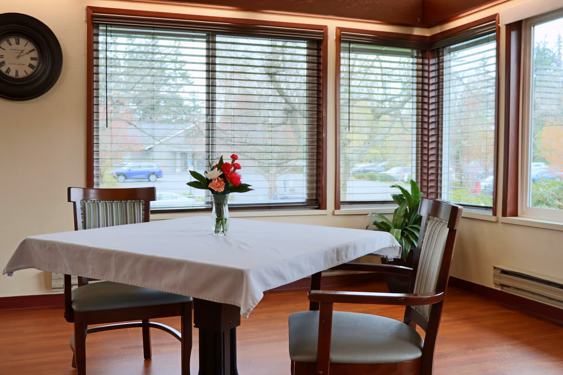 A dining area with a square table covered by a white tablecloth, two wooden chairs with cushions, a vase with red and pink flowers on the table, large windows with wooden blinds showing an outdoor view, a wall clock, and a green plant in the corner.