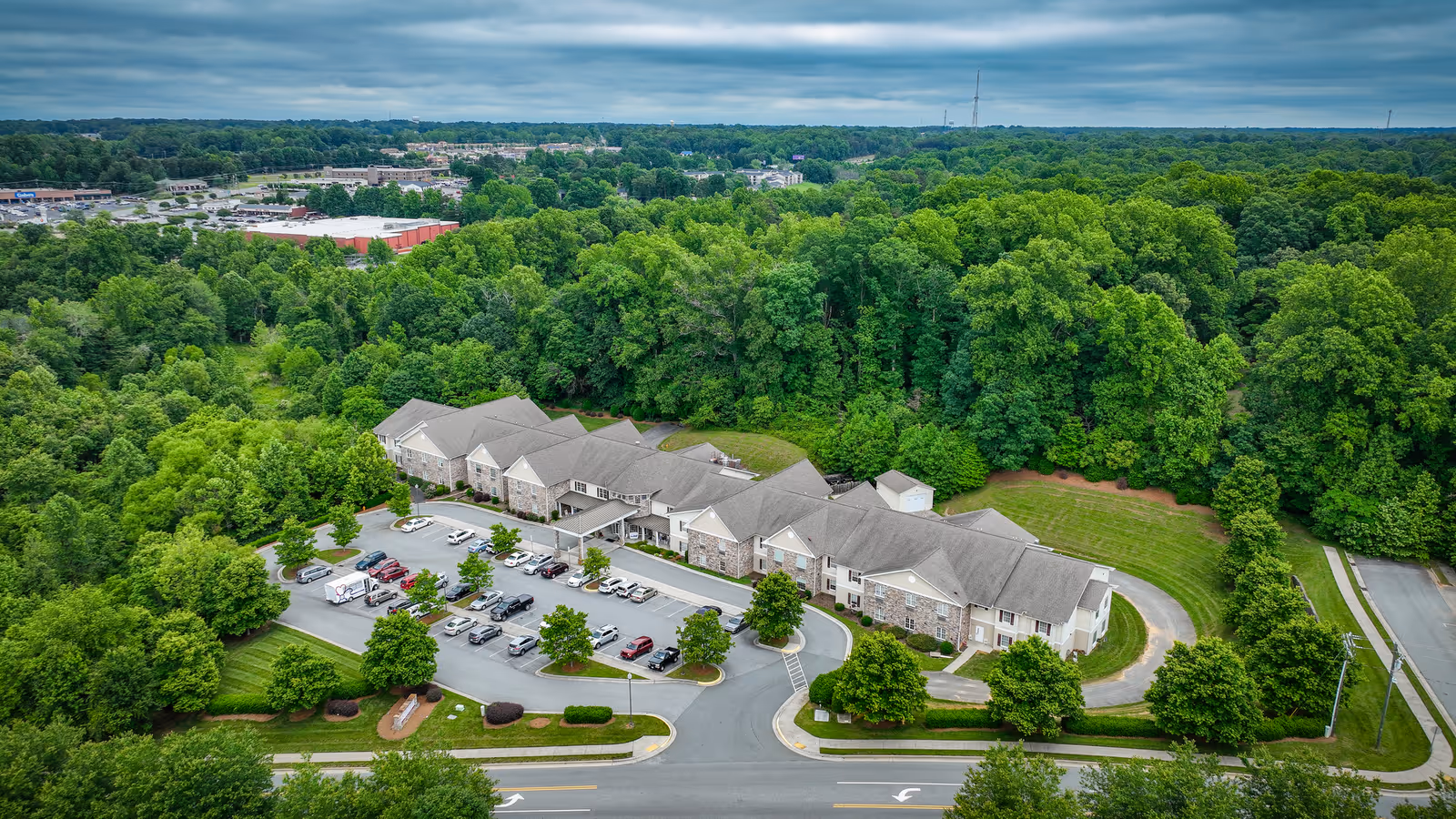 Aerial view of Arbor Ridge At Kernersville, a large senior living facility surrounded by dense green trees. The building has a gray roof and stone facade, with a parking lot filled with cars in front. The facility is situated near a road with landscaped greenery and sidewalks.