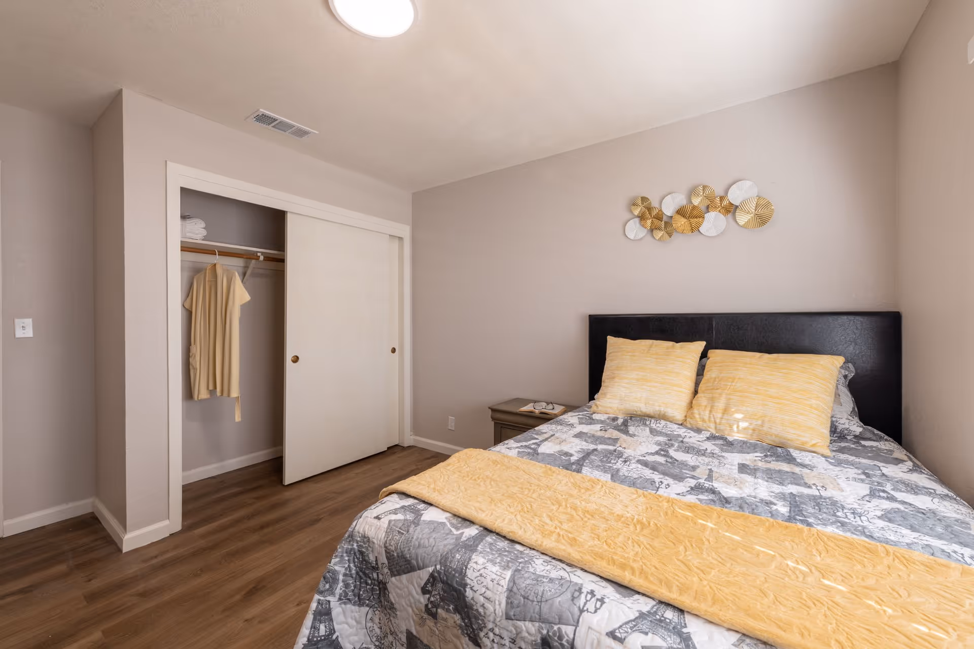 A bedroom in an assisted living facility featuring a bed with a black headboard, two yellow pillows, and a patterned bedspread. There is a yellow throw blanket at the foot of the bed. A nightstand with a telephone is beside the bed. The room has wood flooring, beige walls, and a closet with sliding white doors, one of which is open showing a hanging beige robe and some folded towels on the shelf.