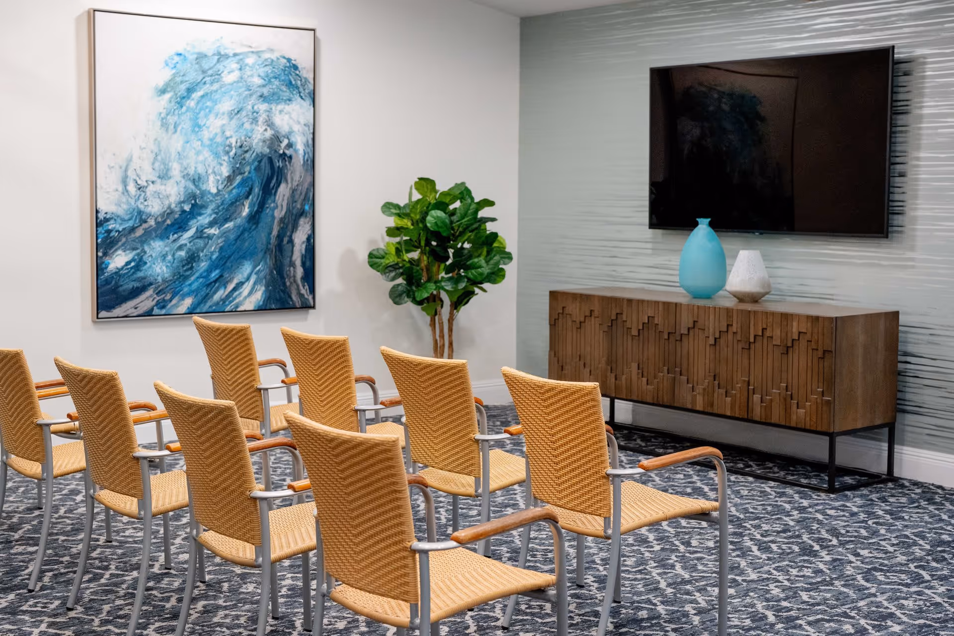 A small meeting or presentation room with two rows of woven chairs facing a wall-mounted flat screen TV. Below the TV is a wooden cabinet with a geometric pattern, decorated with two vases. A large painting of a blue wave and a green potted plant are on the adjacent wall. The room has a patterned carpet and light-colored walls.