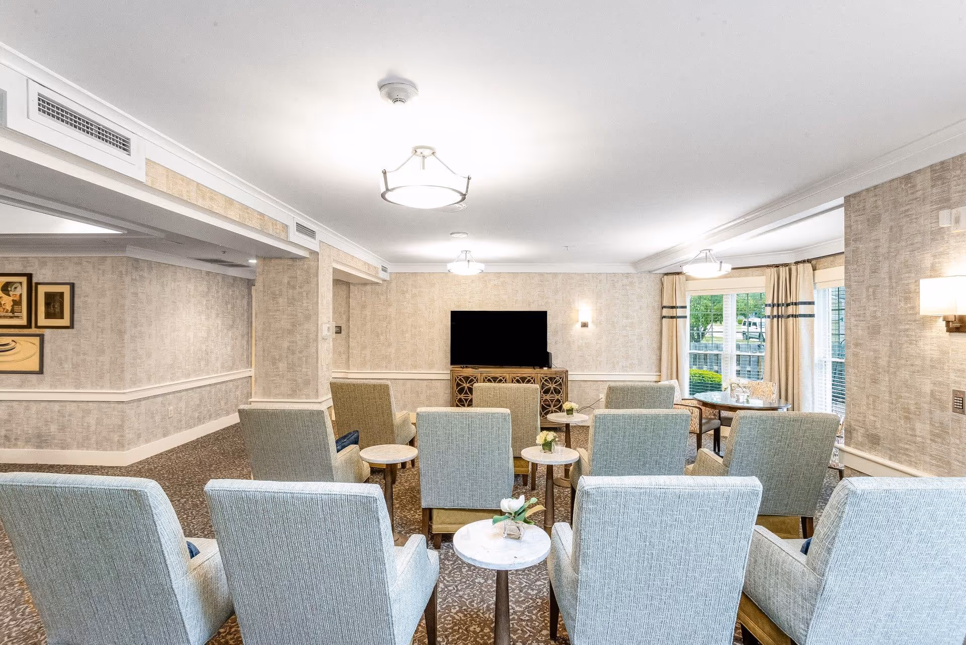 A bright and spacious living room area with multiple light blue upholstered armchairs arranged in rows facing a flat-screen TV mounted on a decorative wooden console. The room features beige patterned wallpaper, carpeted floors, ceiling lights, and large windows with beige curtains letting in natural light. Small round tables with flower arrangements are placed between the chairs.