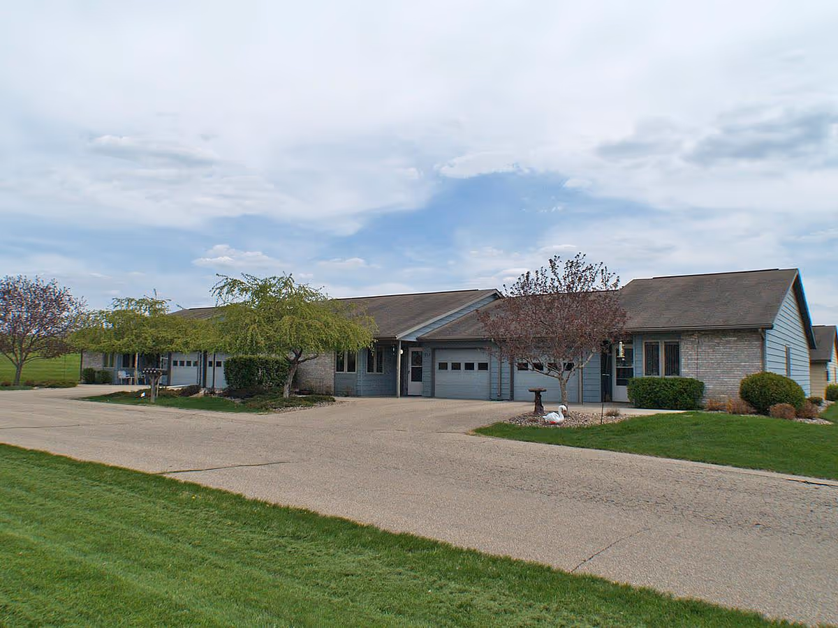 Exterior view of a single-story residential building with attached garages, surrounded by a paved driveway, green lawn, and a few trees under a partly cloudy sky.