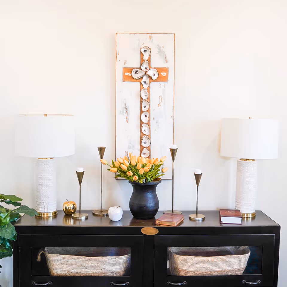 A decorative interior setup featuring a black cabinet with two woven baskets inside. On top of the cabinet are two white table lamps with gold accents, a black vase with orange tulips, several candle holders, two small decorative apples, and two closed books. Above the cabinet hangs a rustic wall art piece with a cross made of oyster shells on a distressed white and orange background.