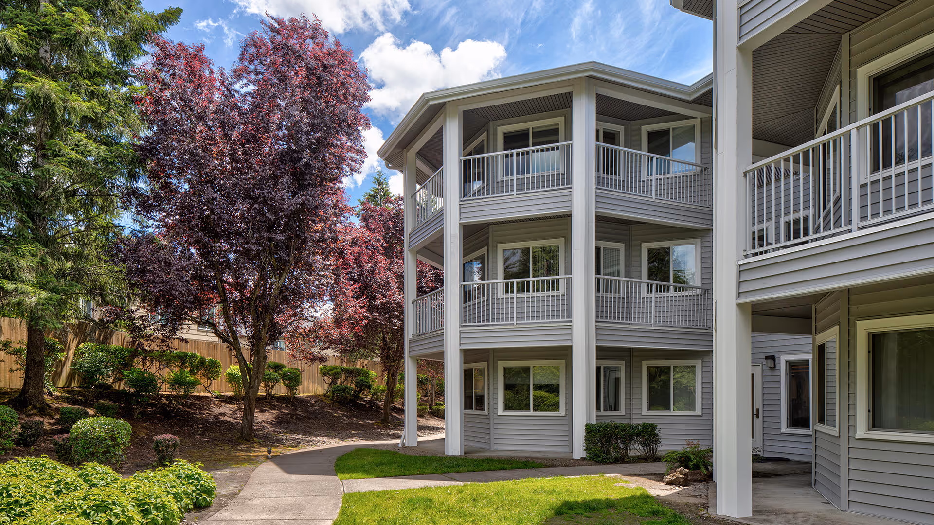 Exterior view of a multi-story residential building with gray siding and white railings on balconies. The building is surrounded by a landscaped garden with green shrubs, trees with red leaves, and a paved walkway under a partly cloudy blue sky.