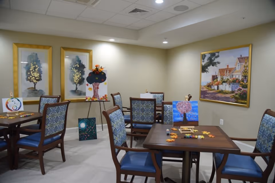A well-lit room with several wooden tables and chairs featuring blue patterned upholstery. The tables display various colorful paintings and art supplies, suggesting an art activity or craft session. The walls are decorated with framed artwork, including two paintings of trees and a larger painting of a house with a garden.