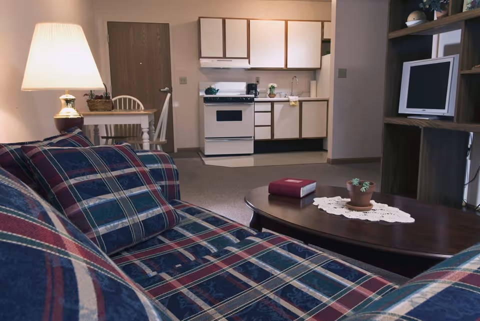 Studio living area with a plaid sofa and coffee table facing a small kitchenette and TV on a wooden shelf.